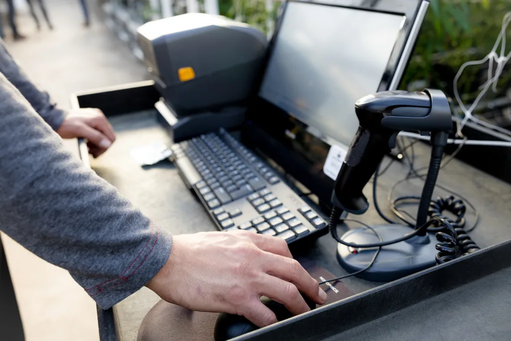Hands at the keyboard and mouse of a weigh-station cart, with a label printer, monitor and handheld scanner in a greenhouse.